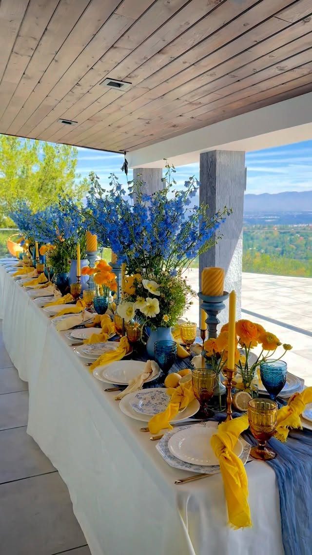 Long dining table decorated with blue flowers, yellow napkins, and blue glassware outdoors.