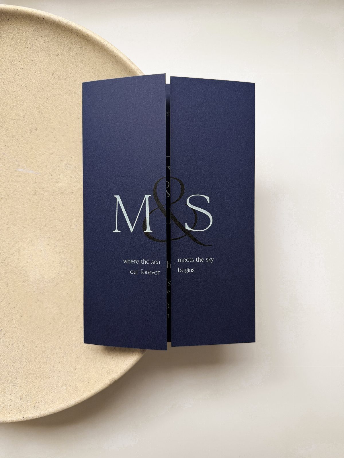 Navy blue wedding invitation with white lettering resting on a beige ceramic plate.