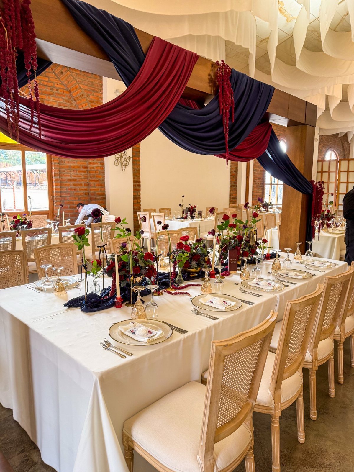 Wedding banquet table with burgundy flowers and navy and burgundy draped fabric overhead.
