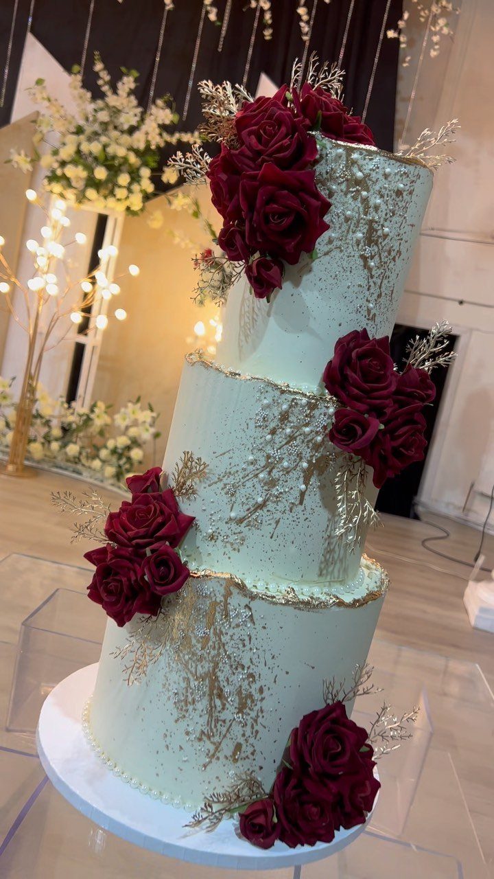 Three-tier white wedding cake with maroon roses and gold leaf metallic accents.