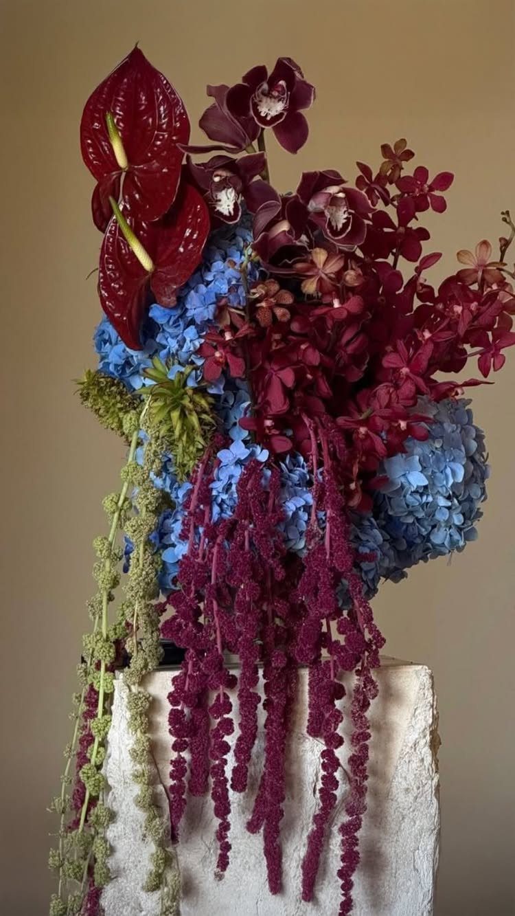 Maroon and blue floral arrangement with hanging amaranthus on a white stone pedestal.
