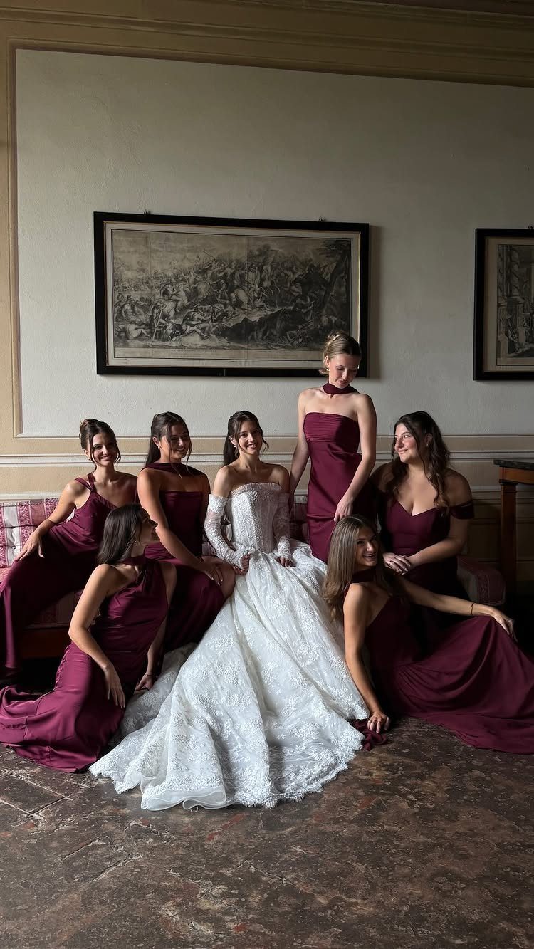 Bride in white lace gown with bridesmaids wearing long maroon dresses posing indoors.