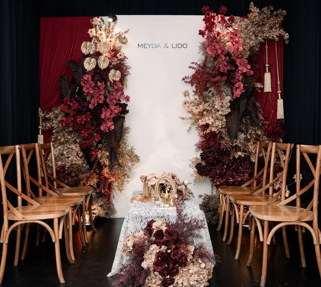 Wedding ceremony backdrop with maroon and gold floral arrangements and wooden cross-back chairs.