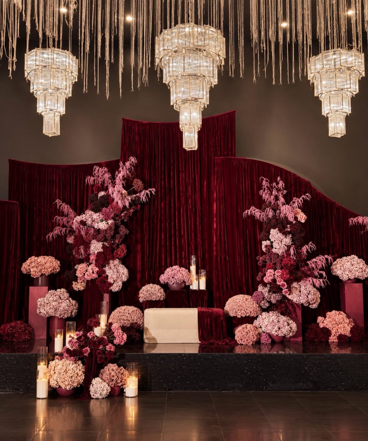 Burgundy and pink floral arrangements set against deep maroon velvet curtains and crystal chandeliers.