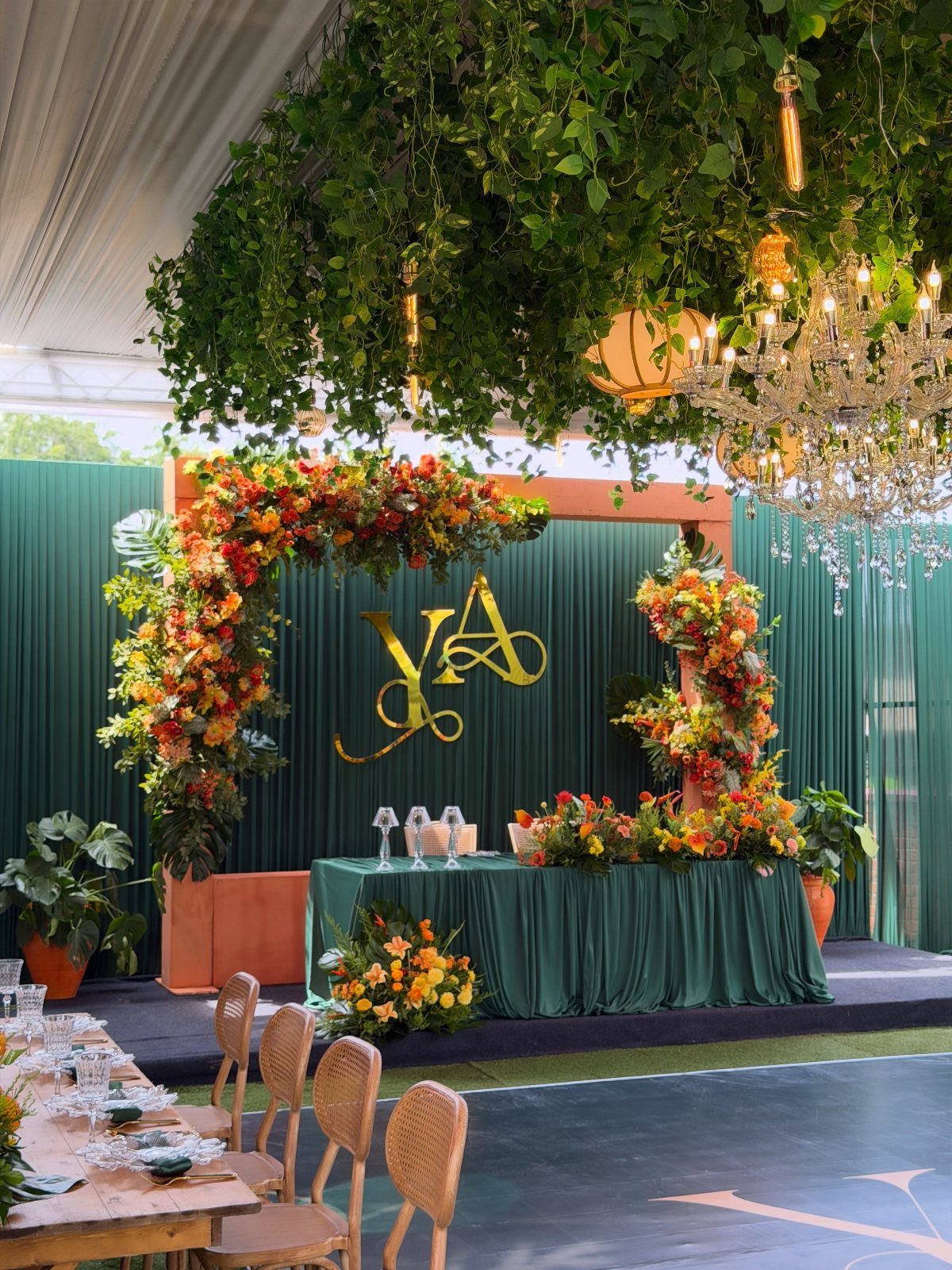 Wedding reception decor with dark green drapes, gold monogram, and orange floral arrangements.