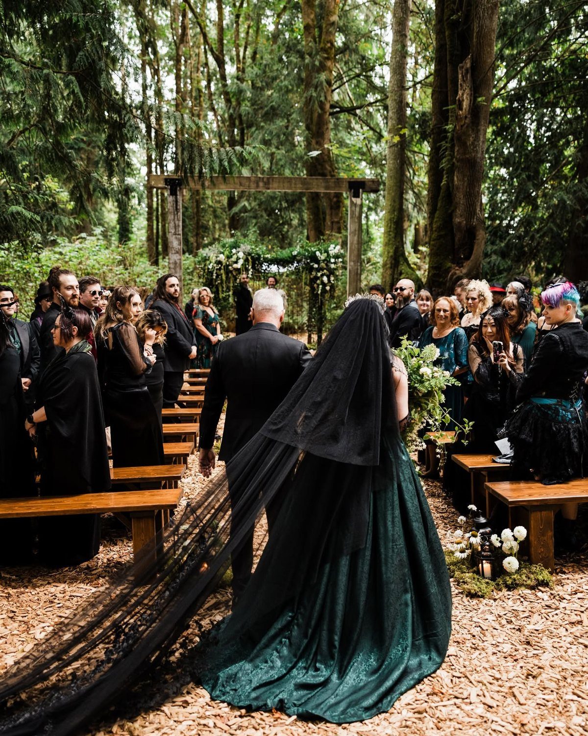 Bride in a dark green wedding gown and black veil walking through woods.