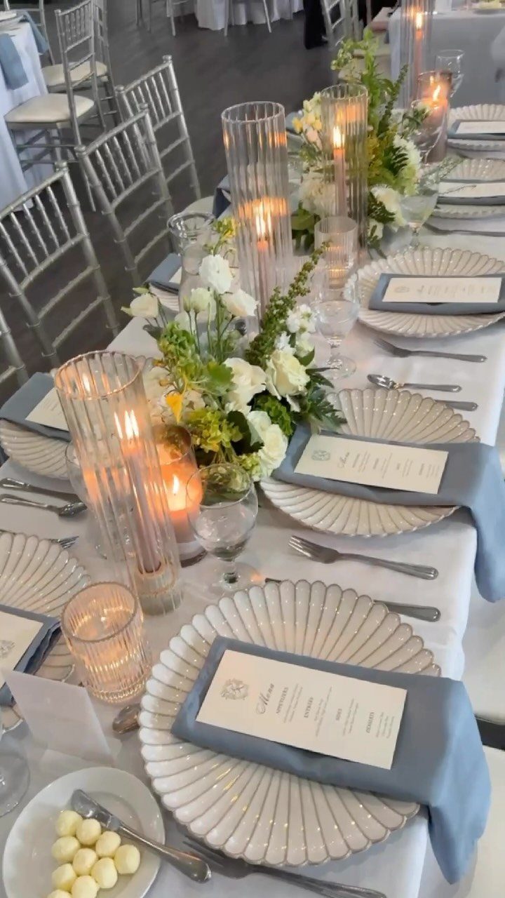 Wedding table setting with dusty blue napkins, white plates, and sage greenery centerpieces.