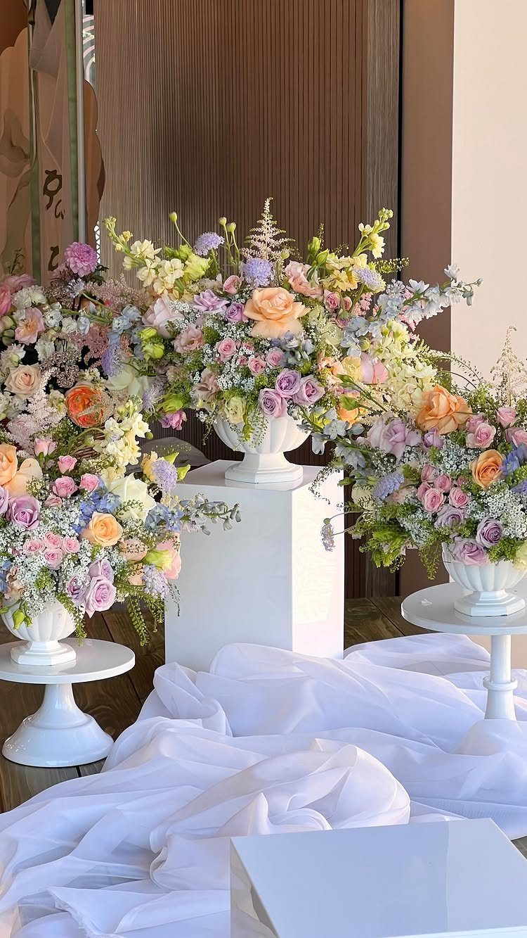 Pastel floral arrangements in white vases on pedestals with draped white fabric.