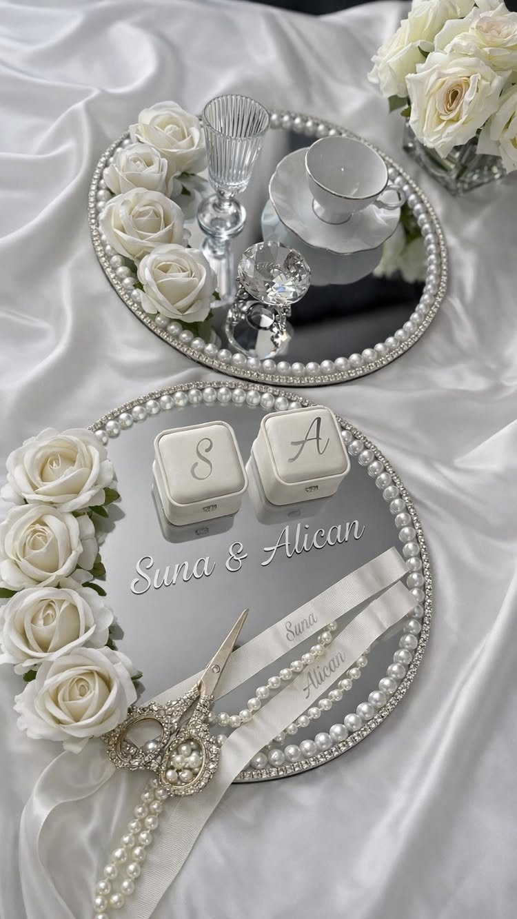 Mirrored trays with pearl edges, white roses, and monogrammed ring boxes on satin fabric.