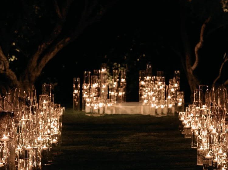 Glowing floating candles in glass cylinders lining a dark outdoor wedding aisle