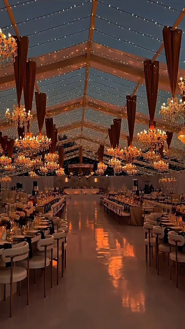 Ornate gold chandeliers hanging from a transparent tent ceiling over wedding reception tables