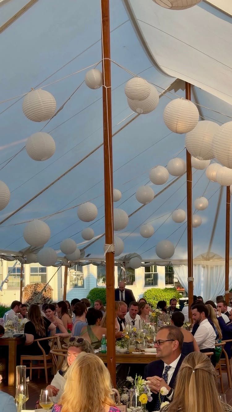 White paper lanterns hanging inside a white tent over wedding guests at dinner.