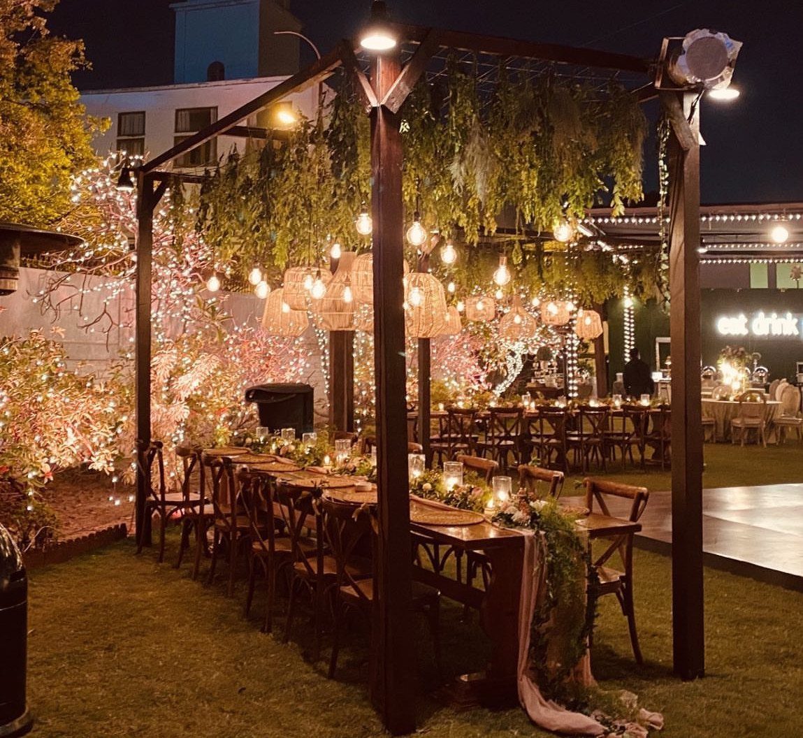 Woven rattan chandeliers hanging over a long wooden wedding table under a pergola.