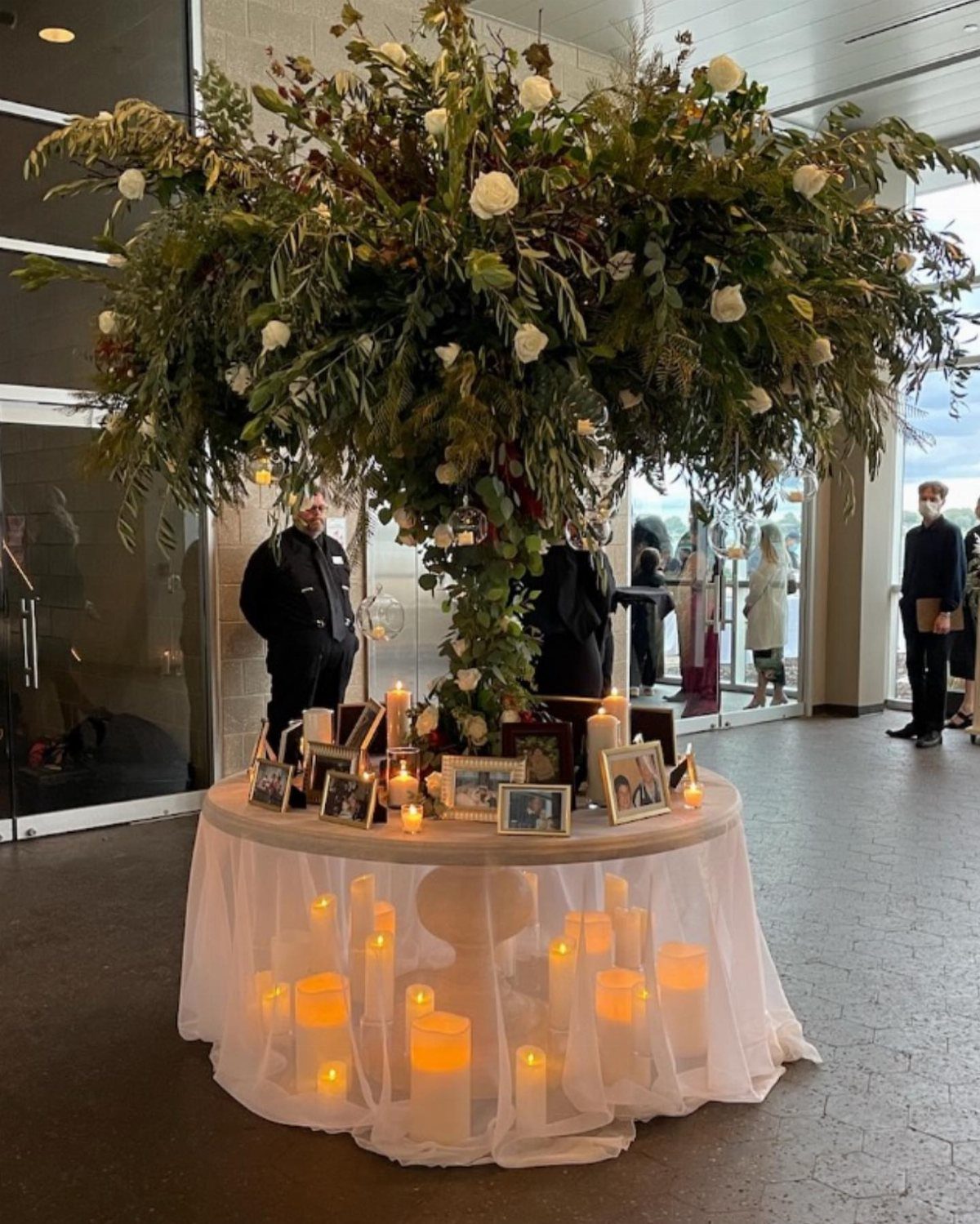 Framed family photographs and candles on a table under a large floral arrangement.