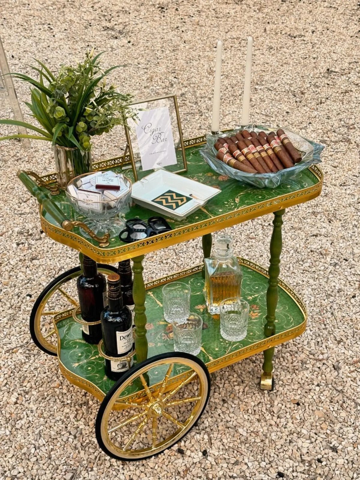 Vintage green and gold bar cart with cigars, liquor, and floral arrangements outdoors.
