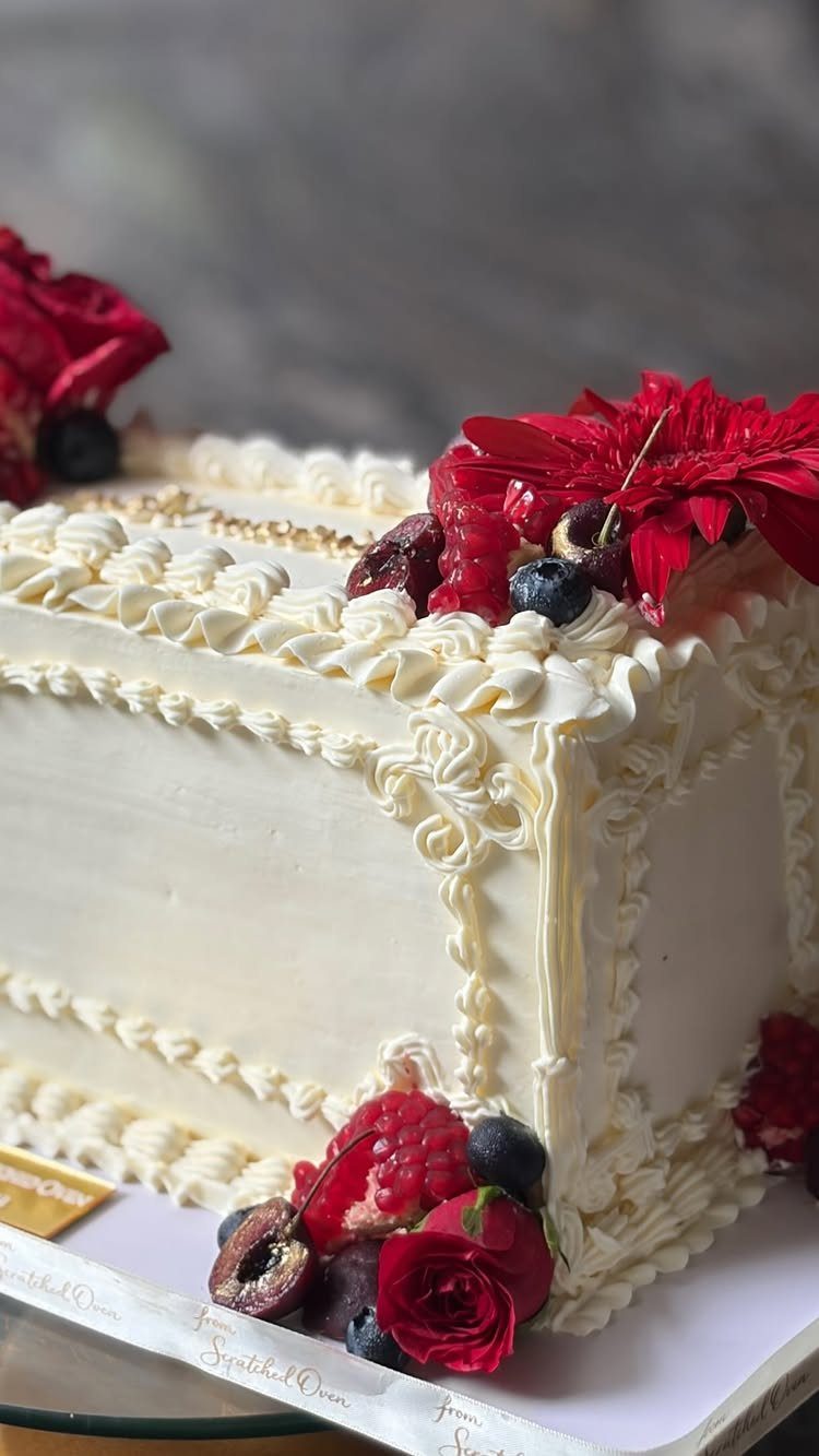 White frosted cake with ornate piping, red roses, and fresh mixed berries.
