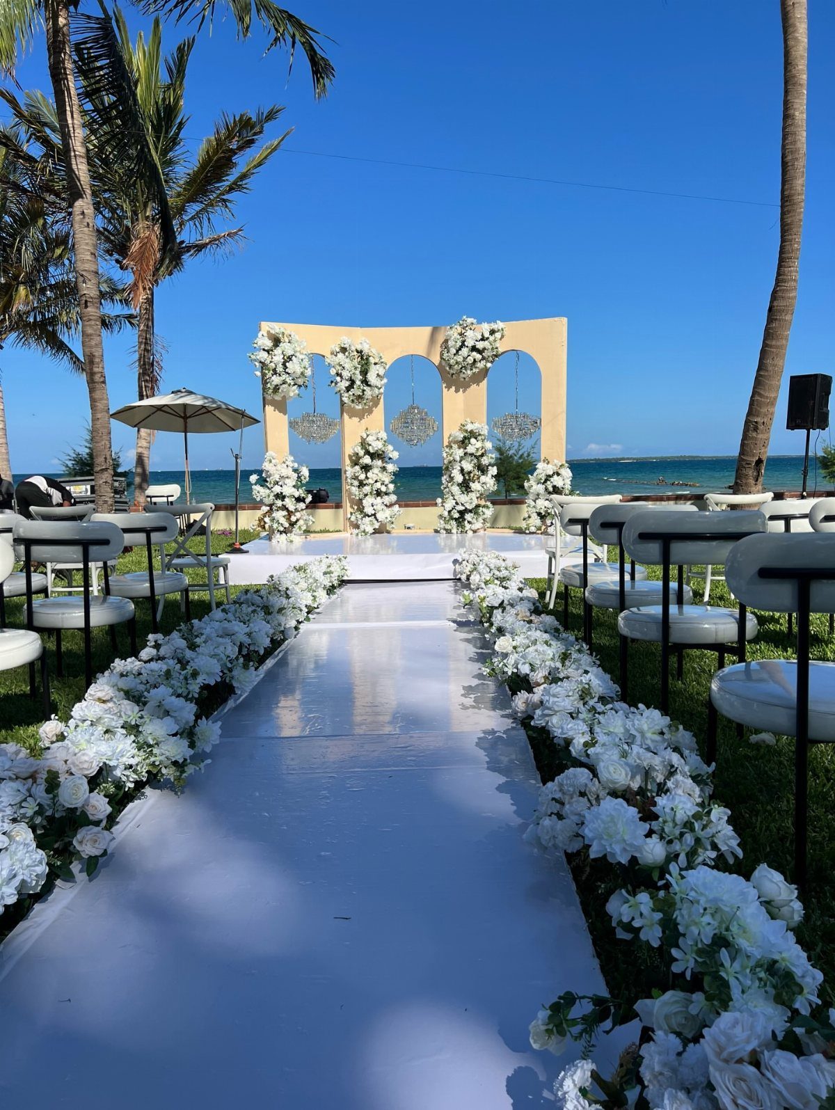 White floral wedding aisle and arched altar on a beach with palm trees.