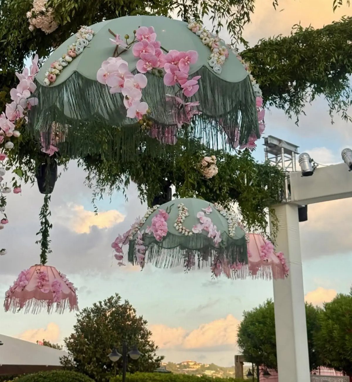 Decorative fringed umbrellas adorned with pink orchids hanging from a leafy tree canopy.