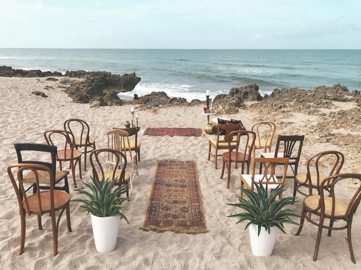 Wooden chairs and patterned rugs arranged for a ceremony on a sandy beach.
