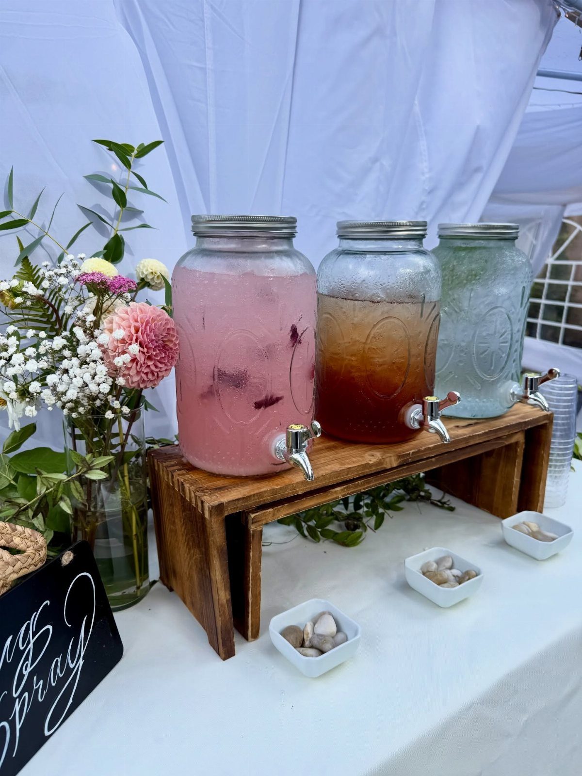 Three beverage dispensers with colorful drinks on a wooden stand with floral accents.