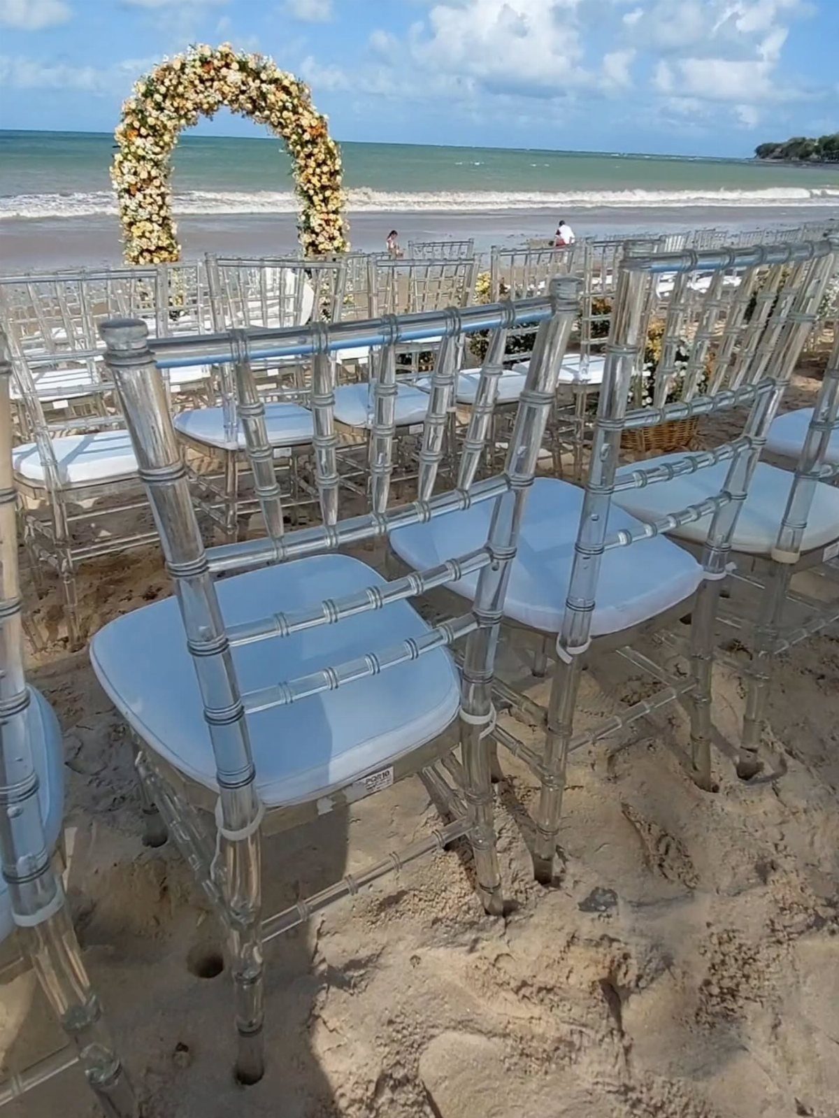 Clear acrylic wedding chairs on a sandy beach facing a floral archway.