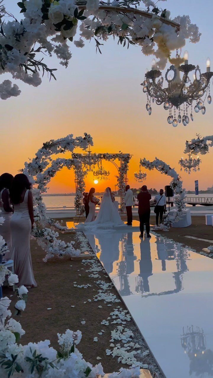 Beach wedding ceremony with white floral arches and chandeliers at sunset