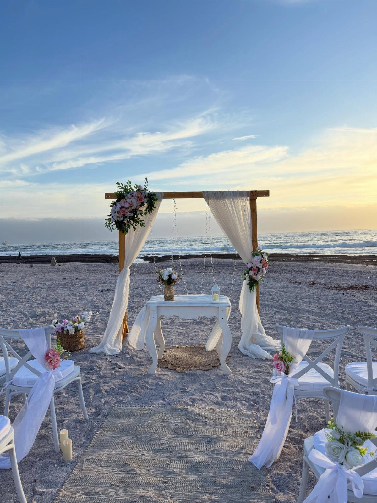 Wooden wedding arbor with white fabric and pink flowers on a sandy beach.
