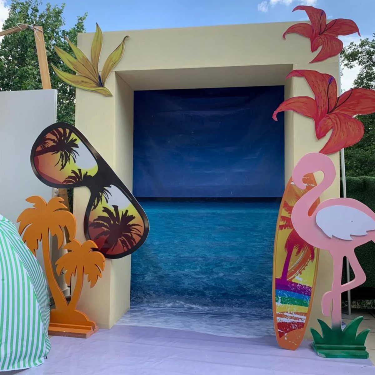 Tropical photo booth setup with ocean backdrop, flamingo, surfboard, and palm tree cutouts.