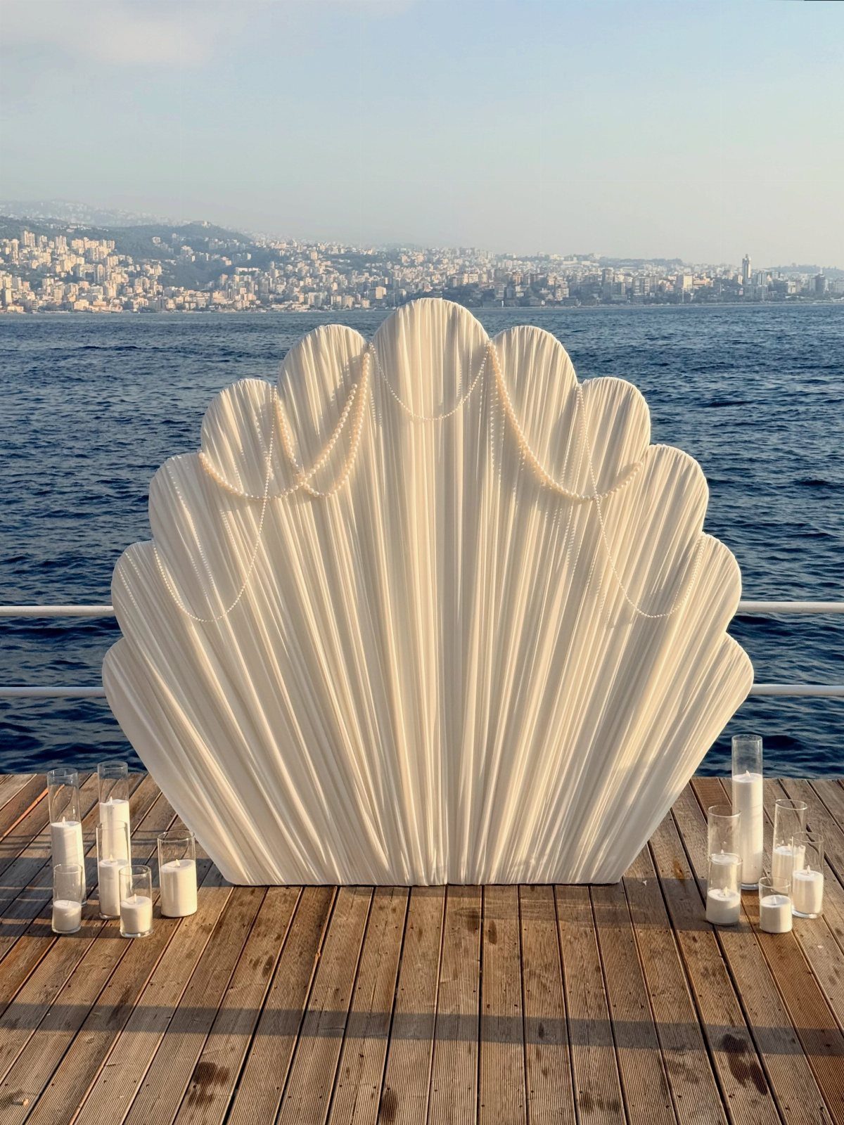 White shell-shaped backdrop with white candles in glass cylinders on a wooden deck