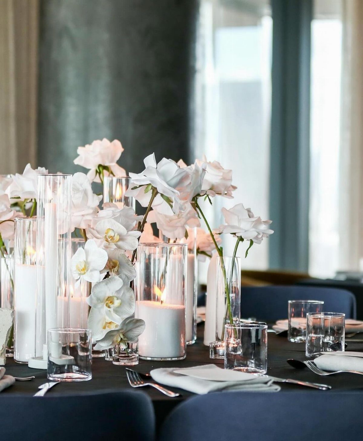 White pillar candles in glass cylinders and white orchids on a dark table.