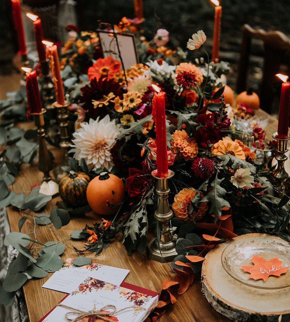 Autumn floral table arrangement with red candles and small orange pumpkins on wood.
