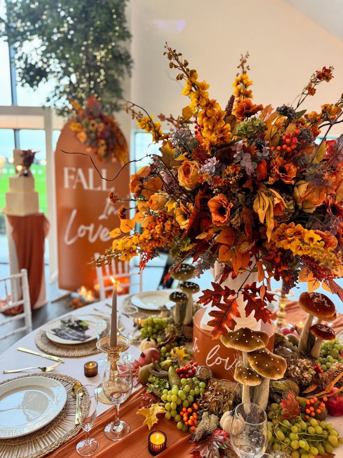 Large autumn floral arrangement with orange blooms, grapes, and mushrooms on a table.