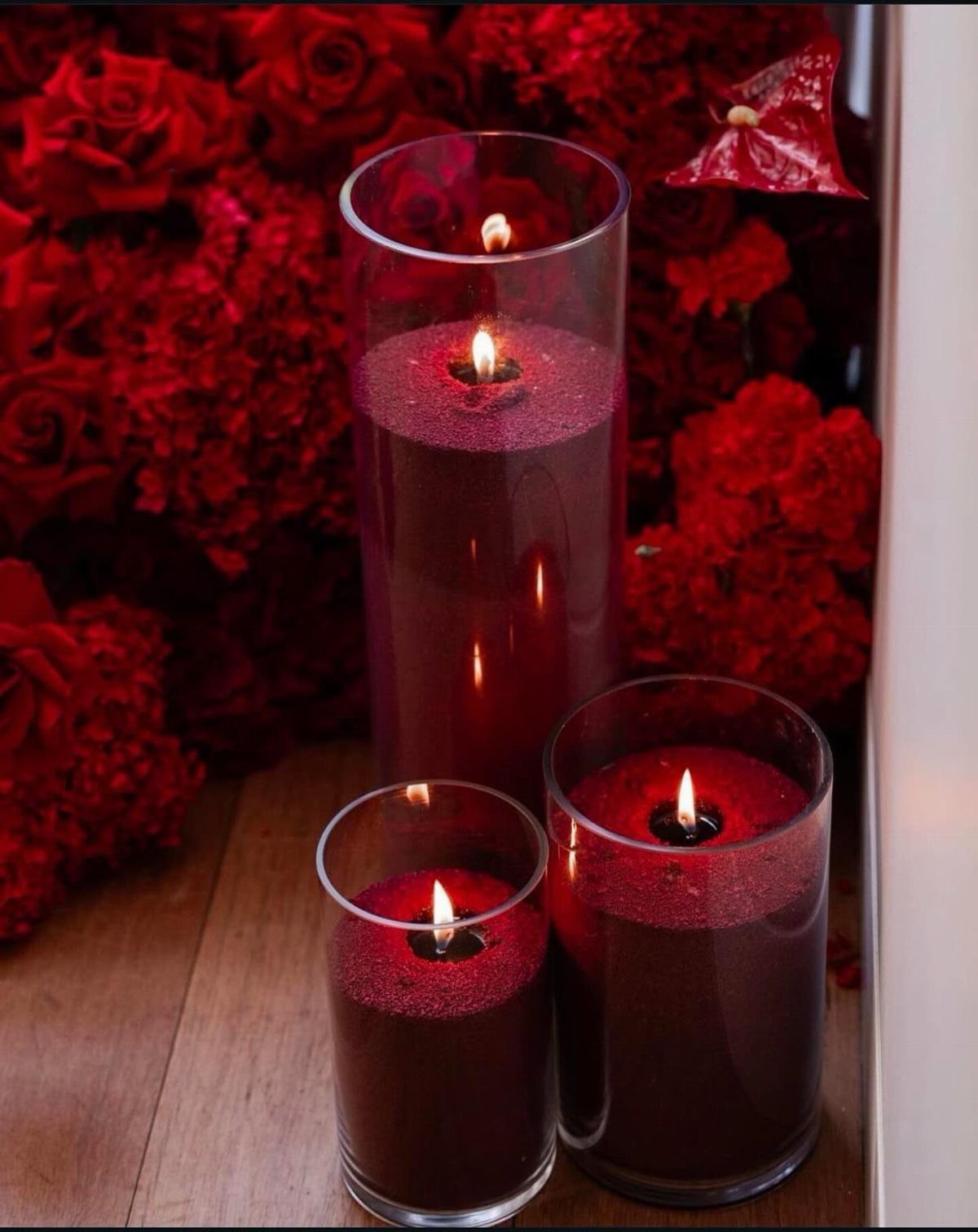 Three lit red candles in glass cylinders against a backdrop of red roses.