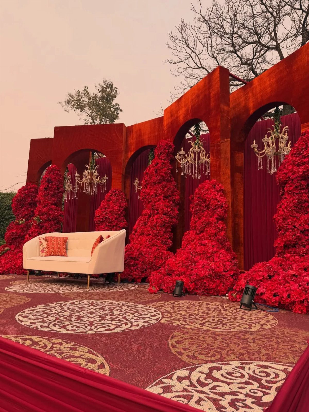 Cream sofa surrounded by red rose mounds and gold chandeliers under red arches.