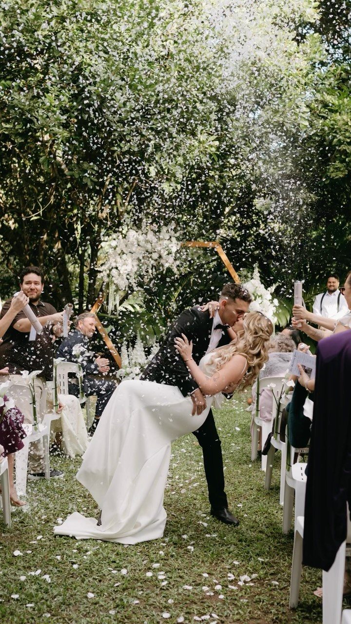 Bride and groom kissing while guests toss white confetti in a garden setting