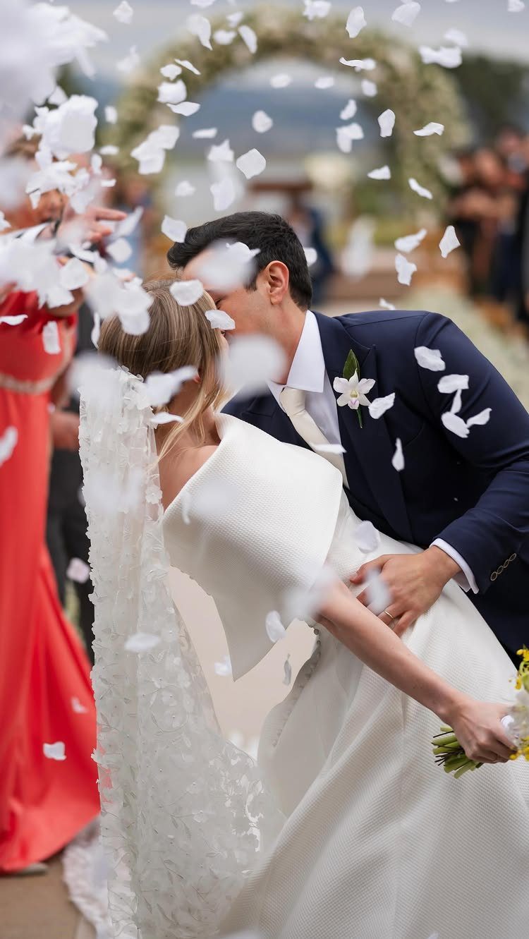 Bride and groom kissing while white flower petals fall around them outdoors
