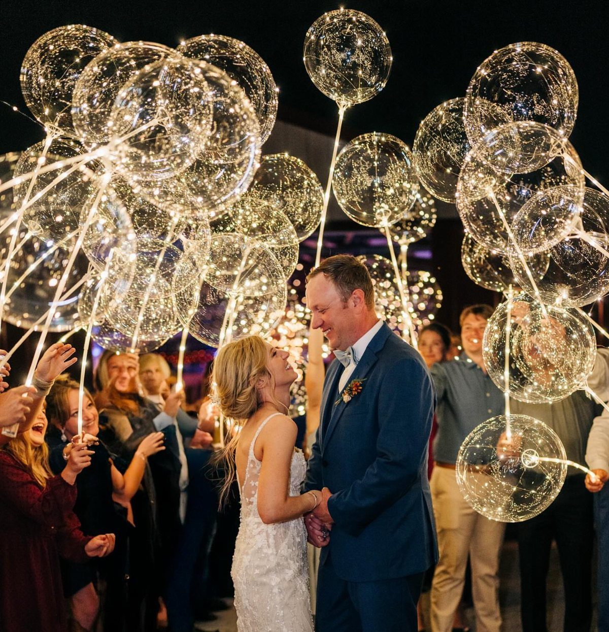 Bride and groom smiling among guests holding glowing LED balloons at night