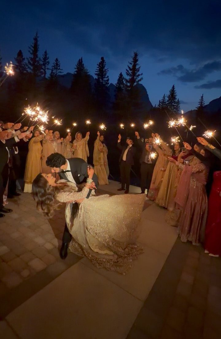Wedding couple kissing while guests hold lit sparklers in a mountain setting at night