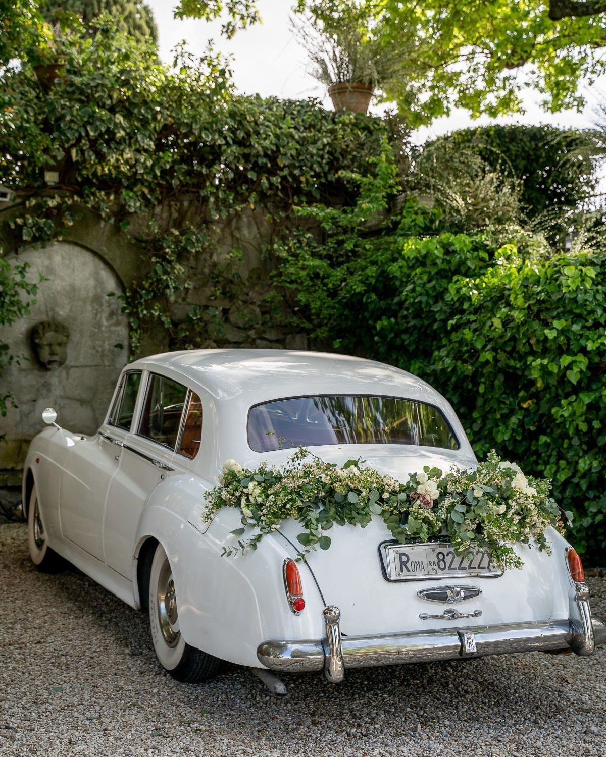 White vintage car with floral decorations on the trunk parked by an ivy wall.