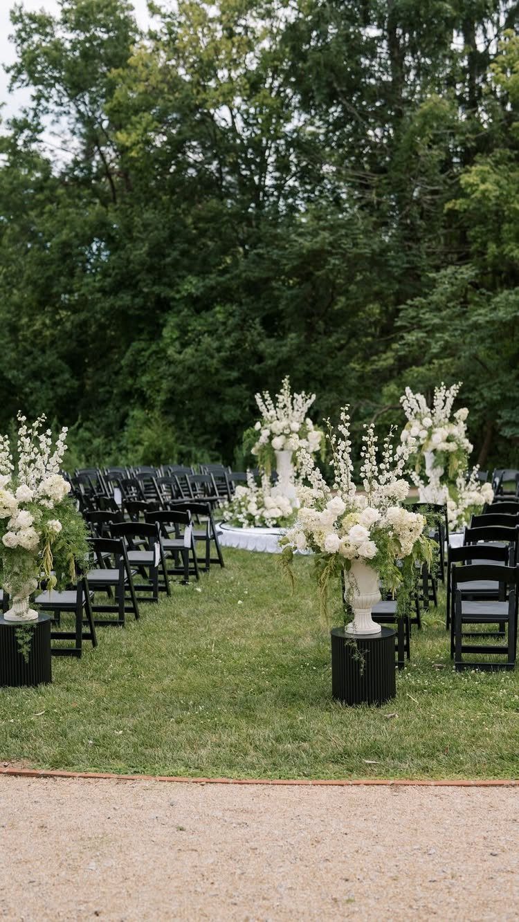 White floral arrangements on black pedestals lining a wedding ceremony aisle outdoors.