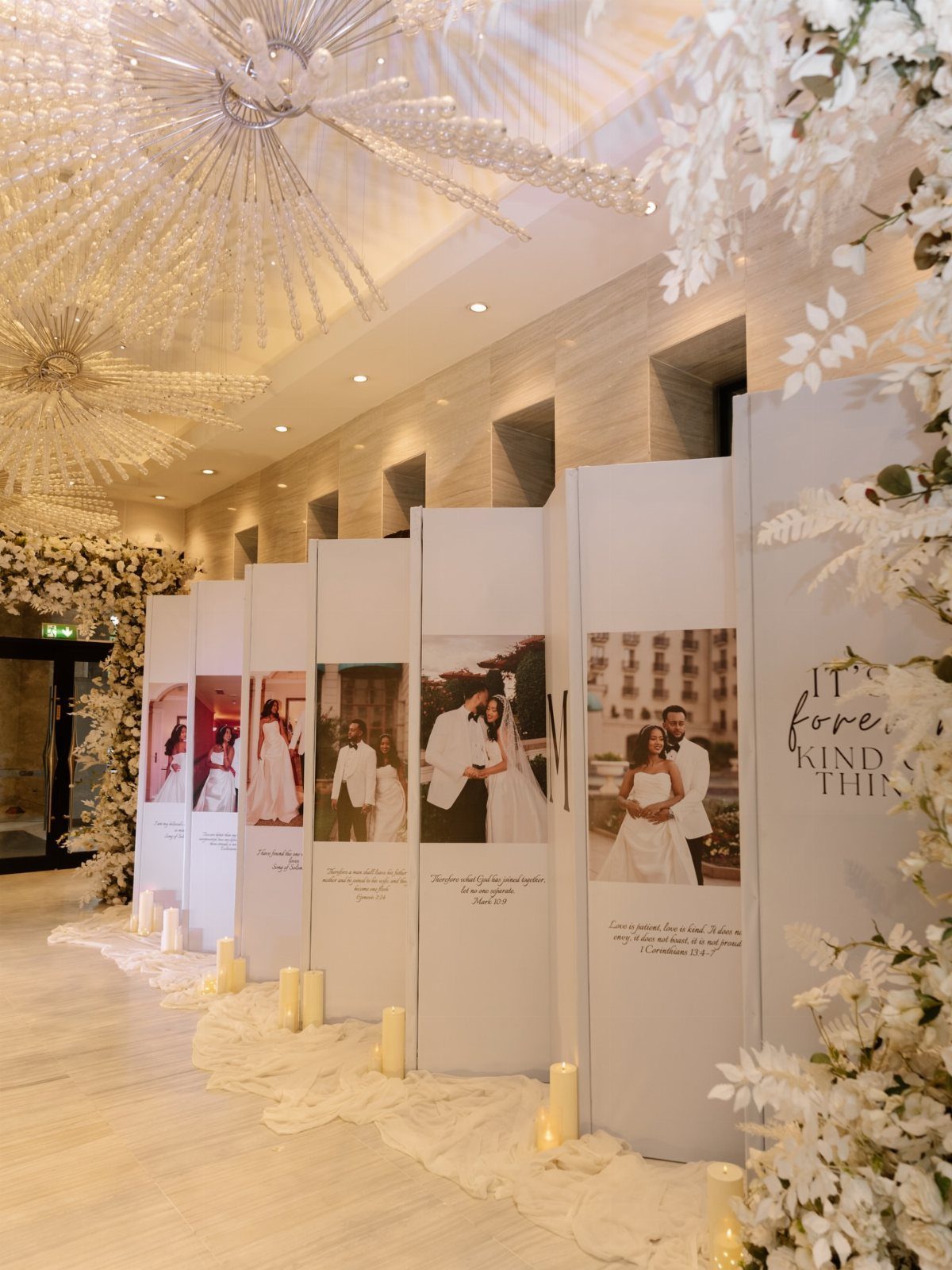White photo panels with wedding pictures, white flowers, and candles in a hall.