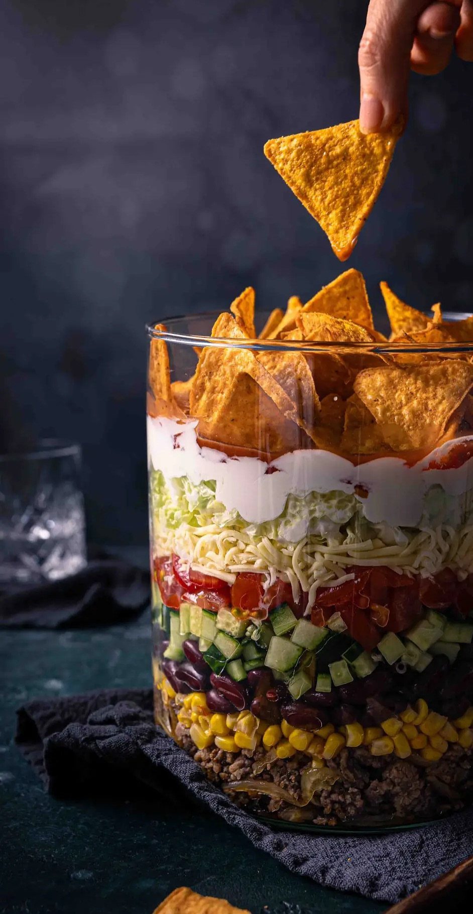 Layered nacho dip in a tall glass cylinder topped with tortilla chips