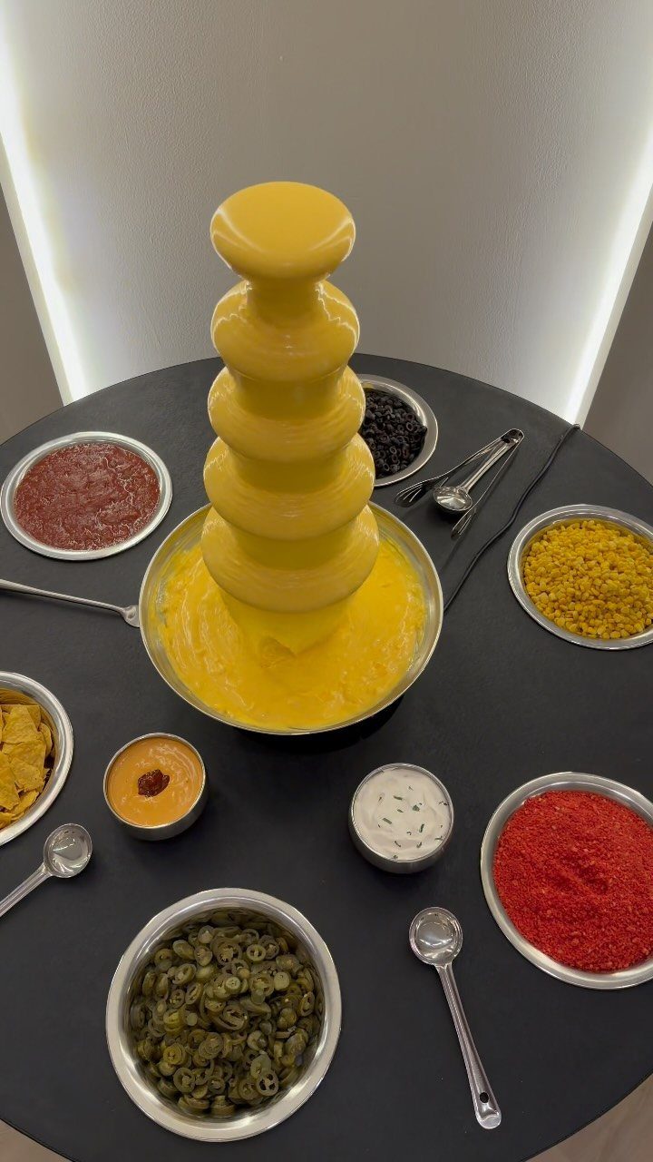 Cheese fountain surrounded by silver bowls of nacho toppings on a black table.