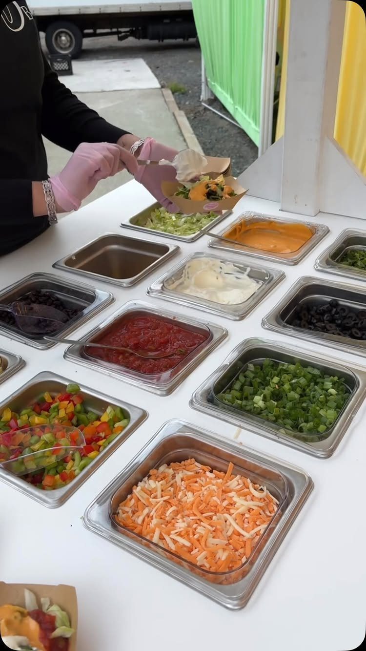 Nacho bar buffet with various toppings in metal trays and a person serving.