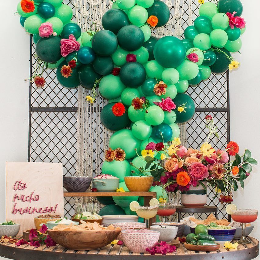 Nacho bar setup with green balloon arch, floral arrangements, and various topping bowls.