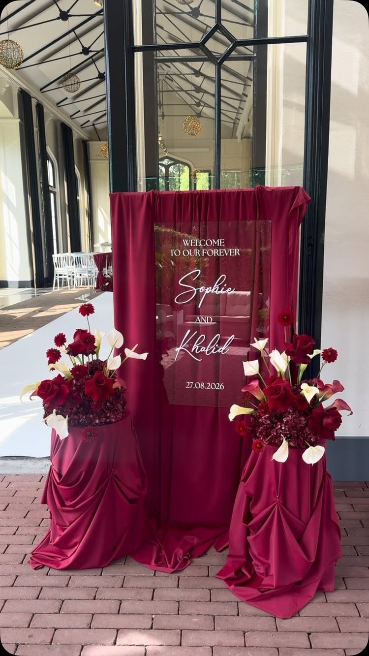 Acrylic welcome sign with red and white floral arrangements and burgundy fabric draping.