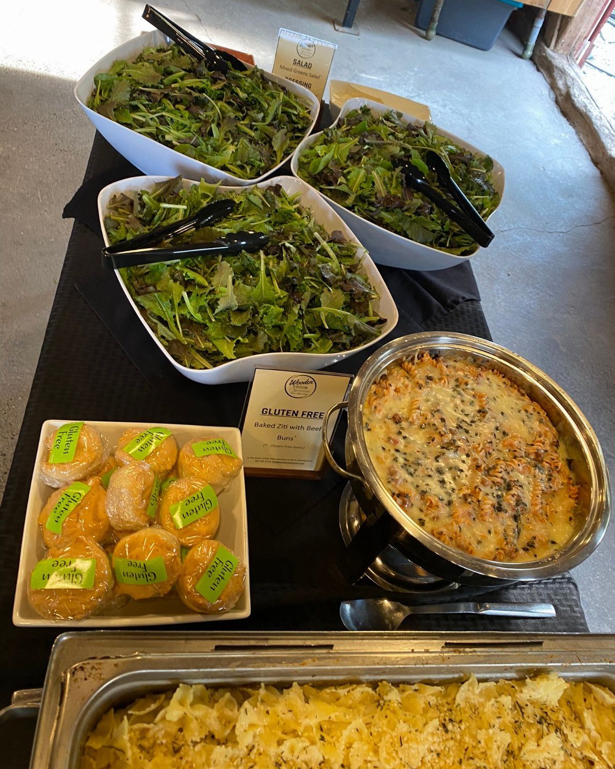 Buffet spread with green salads, baked pasta dishes, and wrapped gluten-free rolls.