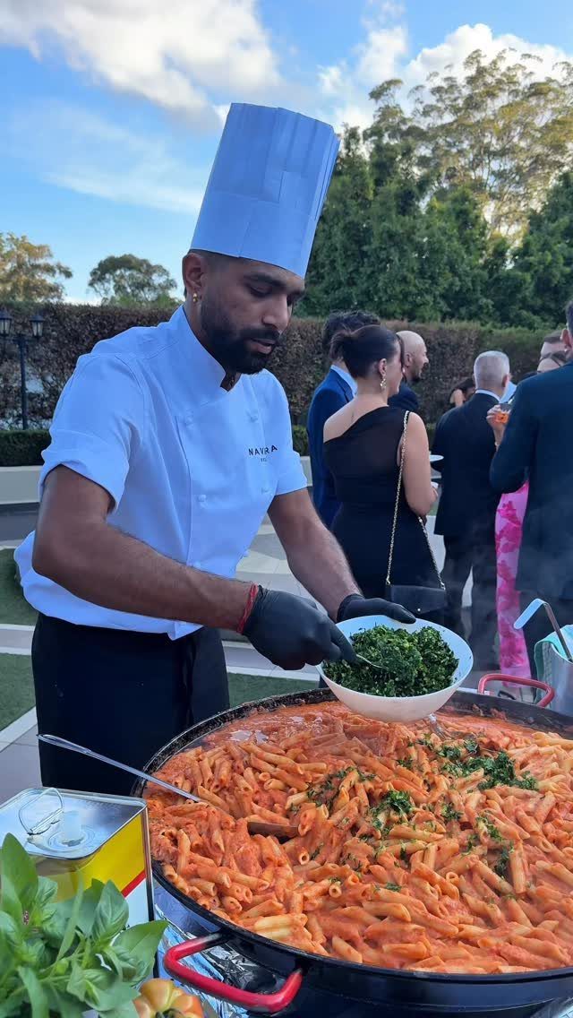 Chef in white uniform preparing penne pasta in a large pan outdoors.