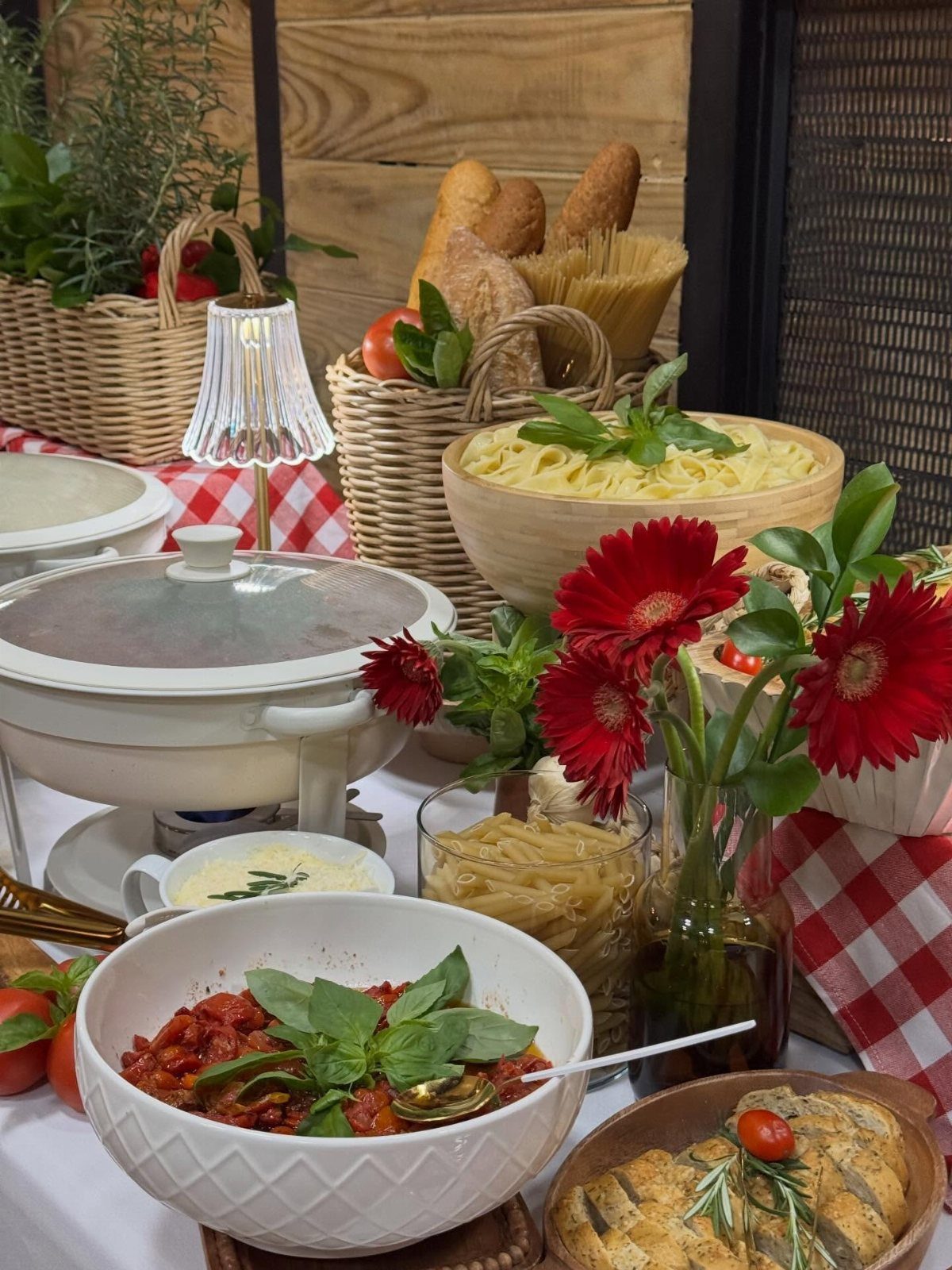 Pasta buffet with red sauce, plain noodles, bread baskets, and red flowers.