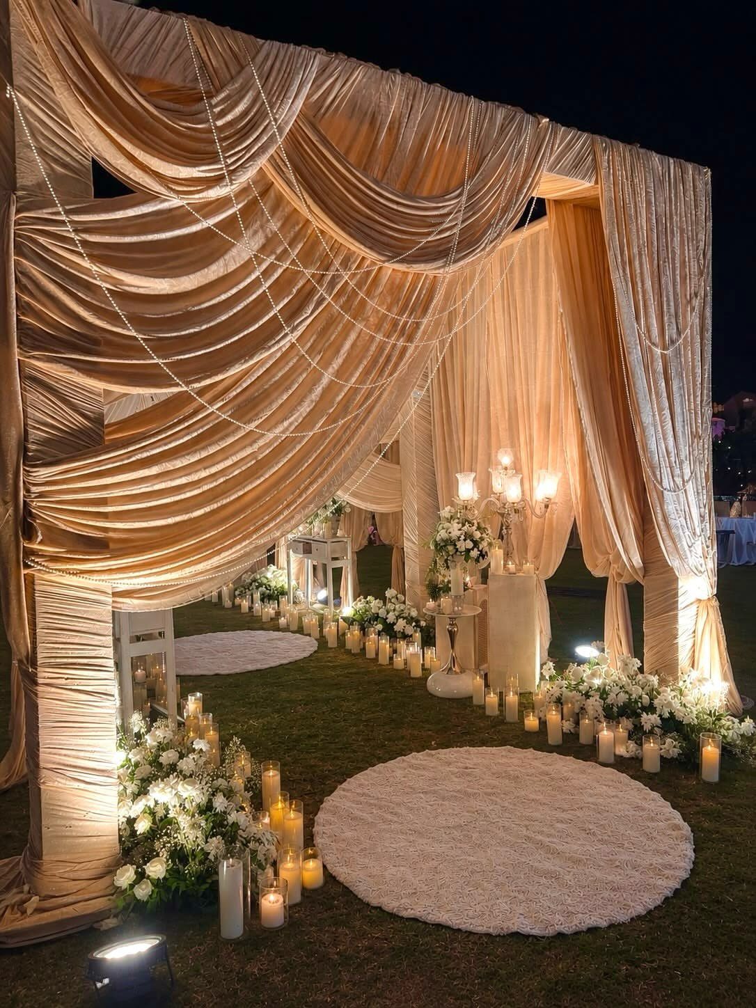 White floral arrangements and pillar candles illuminated by spotlights under draped beige fabric.